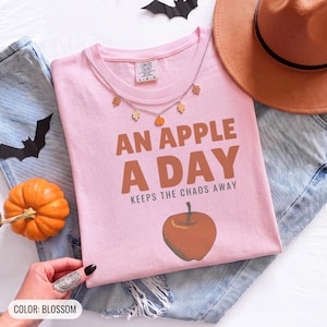 Blossom Comfort Colors t-shirt printed with the words An Apple A Day Keeps The Chaos Away and a drawing of a shiny red apple with a single green leaf and a tiny worm peeking out, worn by a woman