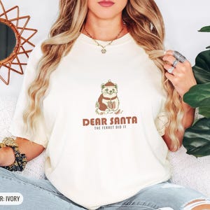 Ivory Comfort Colors t-shirt printed with the words Dear Santa The Ferret Did It and a drawing of a mischievous ferret dragging a striped Christmas stocking filled with shiny ornaments, worn by a woman