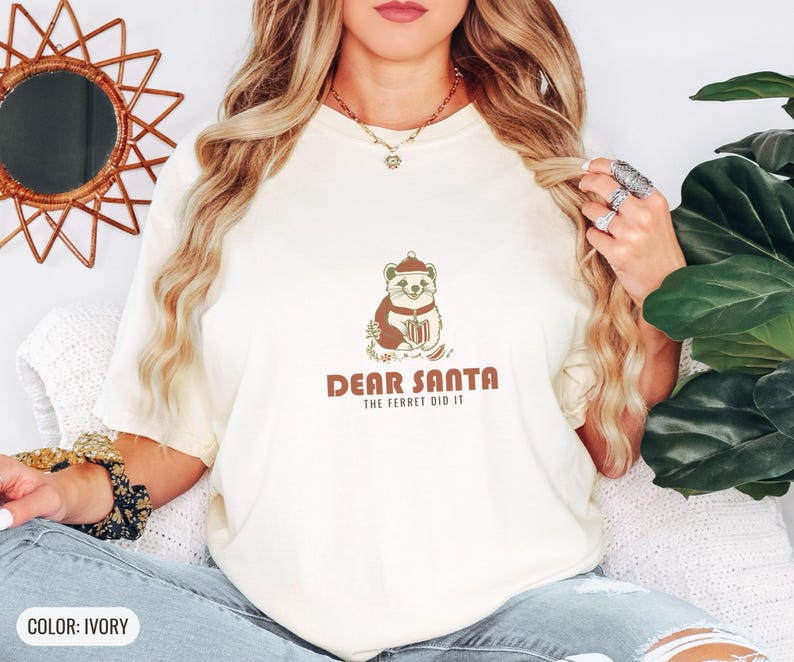 Ivory Comfort Colors t-shirt printed with the words Dear Santa The Ferret Did It and a drawing of a mischievous ferret dragging a striped Christmas stocking filled with shiny ornaments, worn by a woman