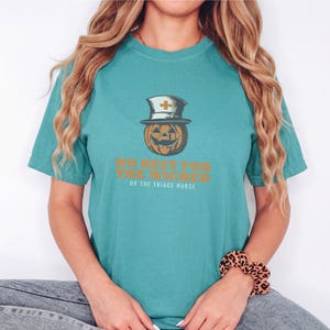 Seafoam Comfort Colors t-shirt printed with the words No Rest For The Wicked Or The Triage Nurse and a drawing of a grinning jack-o'-lantern wearing a classic nurse's cap with a red cross, worn by a woman