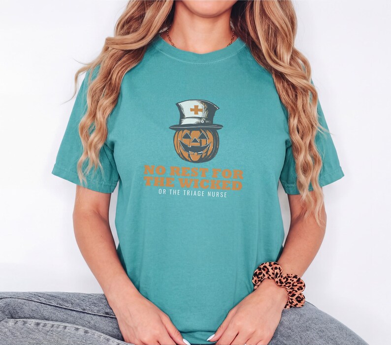 Seafoam Comfort Colors t-shirt printed with the words No Rest For The Wicked Or The Triage Nurse and a drawing of a grinning jack-o'-lantern wearing a classic nurse's cap with a red cross, worn by a woman