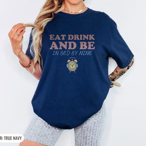 True Navy Comfort Colors t-shirt printed with the words Eat Drink And Be In Bed By Nine and a drawing of a retro brass wind-up alarm clock with its bells wrapped in fuzzy winter earmuffs, worn by a woman