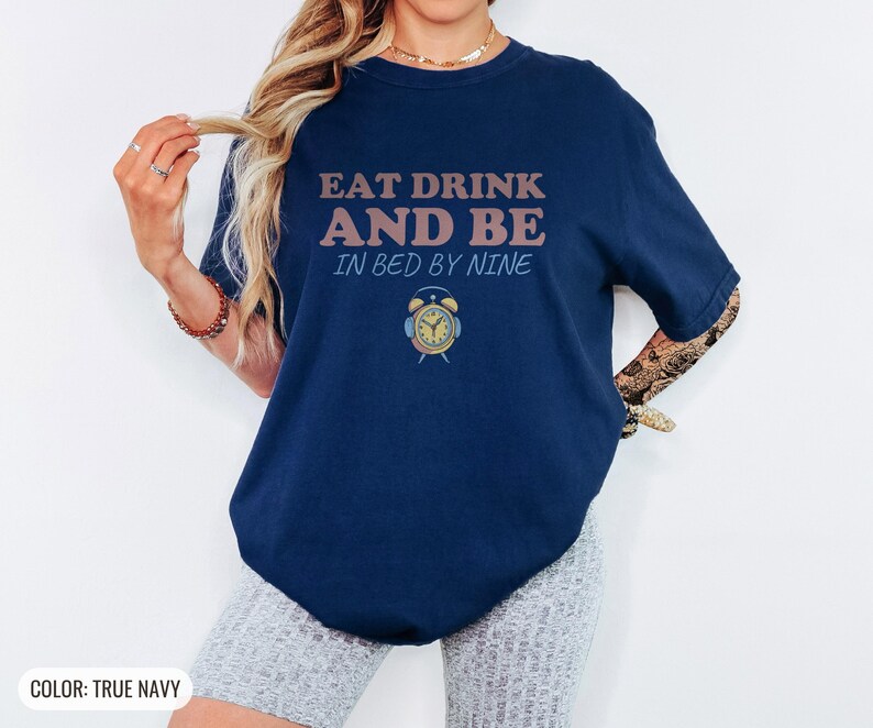True Navy Comfort Colors t-shirt printed with the words Eat Drink And Be In Bed By Nine and a drawing of a retro brass wind-up alarm clock with its bells wrapped in fuzzy winter earmuffs, worn by a woman