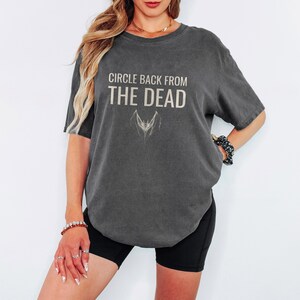 Pepper Comfort Colors t-shirt printed with the words Circle Back From The Dead and a drawing of a vintage silver paperclip bent into the shape of a bat, worn by a woman