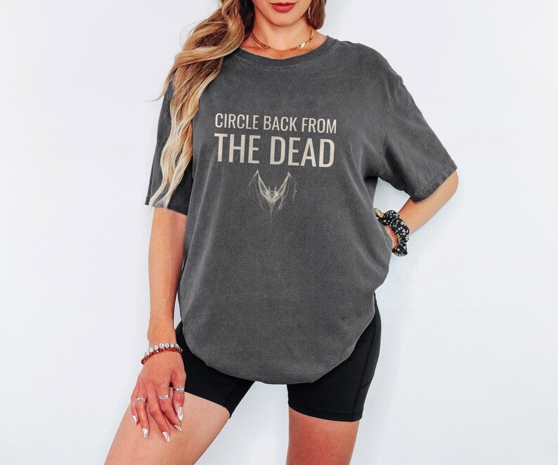 Pepper Comfort Colors t-shirt printed with the words Circle Back From The Dead and a drawing of a vintage silver paperclip bent into the shape of a bat, worn by a woman
