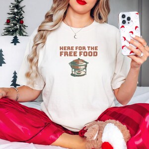 Ivory Comfort Colors t-shirt printed with the words Here For The Free Food and a drawing of a stainless steel chafing dish with steam rising off the lid, worn by a woman