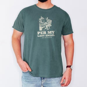 Blue Spruce Comfort Colors t-shirt printed with the words Per My Last Email I Am Deceased and a drawing of a skeleton sitting at an office desk typing on a computer keyboard, worn by a man