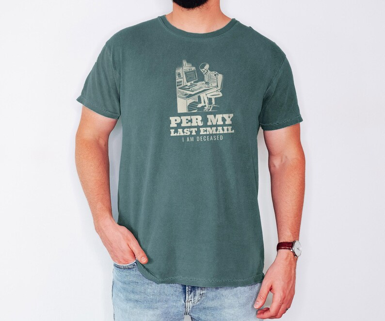 Blue Spruce Comfort Colors t-shirt printed with the words Per My Last Email I Am Deceased and a drawing of a skeleton sitting at an office desk typing on a computer keyboard, worn by a man