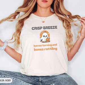 Ivory Comfort Colors t-shirt printed with the words Crisp Breeze Leaves Turning And Bones Rattling and a drawing of a large cute smiling ghost beside a carved jack-o-lantern, worn by a woman