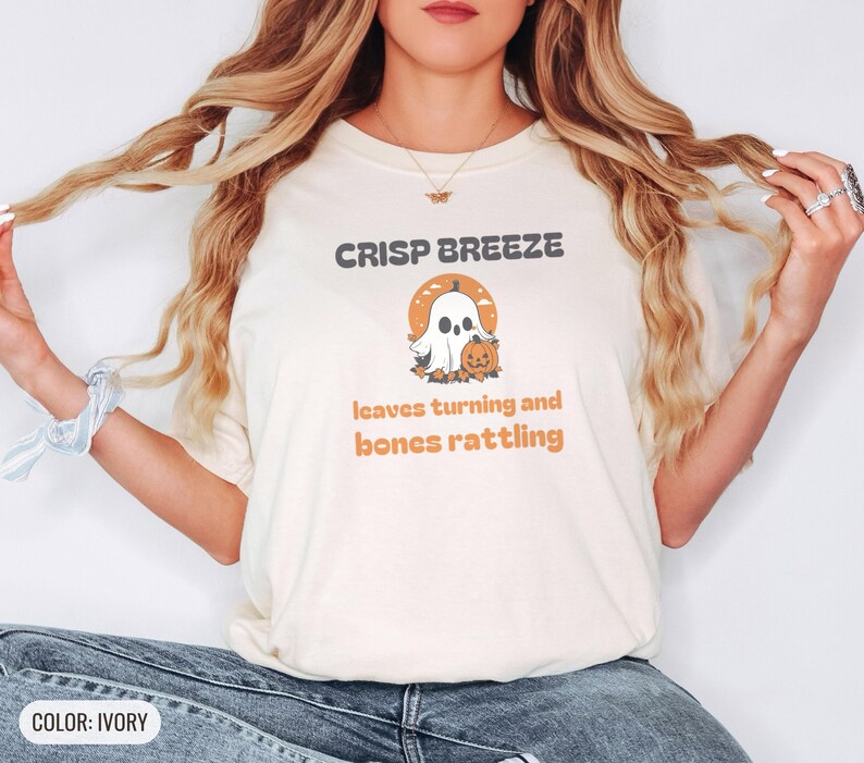 Ivory Comfort Colors t-shirt printed with the words Crisp Breeze Leaves Turning And Bones Rattling and a drawing of a large cute smiling ghost beside a carved jack-o-lantern, worn by a woman
