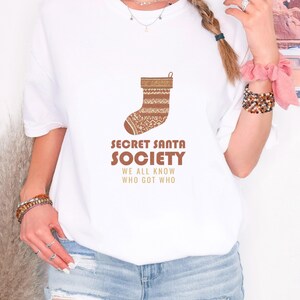 White Comfort Colors t-shirt printed with the words Secret Santa Society We All Know Who Got Who and a drawing of a knitted wool stocking stuffed with stick candy, worn by a woman