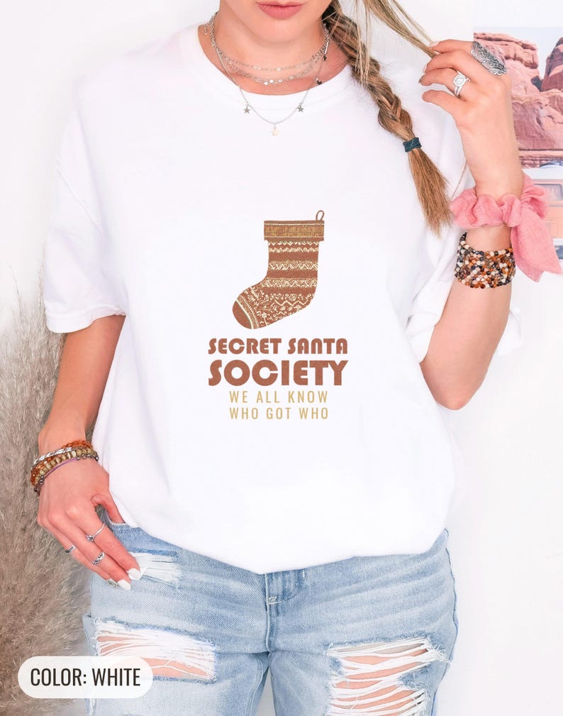 White Comfort Colors t-shirt printed with the words Secret Santa Society We All Know Who Got Who and a drawing of a knitted wool stocking stuffed with stick candy, worn by a woman