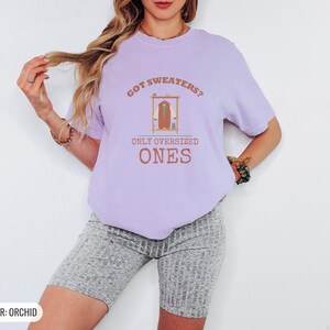 Orchid Comfort Colors t-shirt printed with the words Got Sweaters? Only Oversized Ones and a drawing of a knitted wool cardigan hanging from a simple wooden clothes hanger, worn by a woman