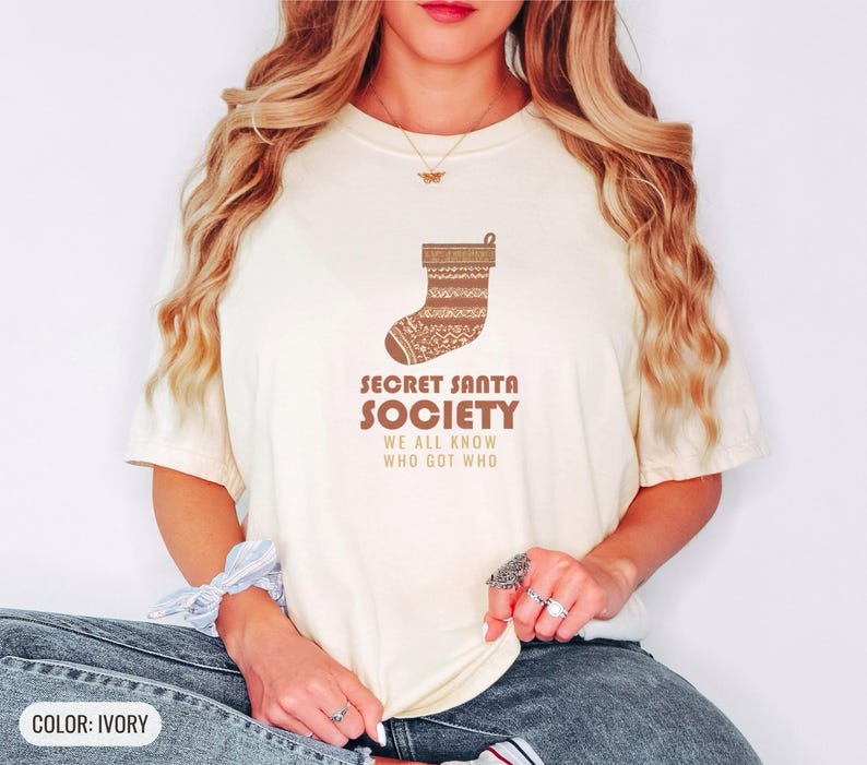 Ivory Comfort Colors t-shirt printed with the words Secret Santa Society We All Know Who Got Who and a drawing of a knitted wool stocking stuffed with stick candy, worn by a woman