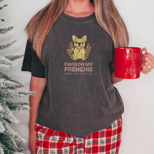 Pepper Comfort Colors t-shirt printed with the words Pardon My Frenchie Under The Mistletoe and a drawing of a vintage brass French Bulldog bottle opener lying next to a sprig of red-berried mistleto, worn by a woman
