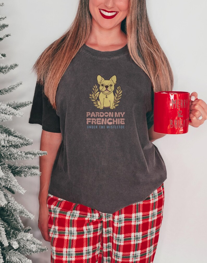 Pepper Comfort Colors t-shirt printed with the words Pardon My Frenchie Under The Mistletoe and a drawing of a vintage brass French Bulldog bottle opener lying next to a sprig of red-berried mistleto, worn by a woman