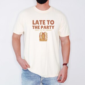 Ivory Comfort Colors t-shirt printed with the words Late To The Party Early To The Library and a drawing of a woodland owl wearing spectacles sitting on a book, worn by a man