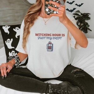 Ivory Comfort Colors t-shirt printed with the words Witching Hour Is Just My Shift and a drawing of a retro medical defibrillator machine decorated with a spooky jack-o-lantern face on its m, worn by a woman