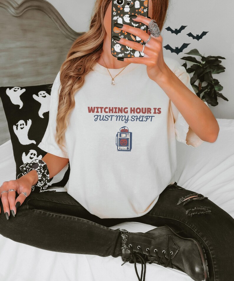 Ivory Comfort Colors t-shirt printed with the words Witching Hour Is Just My Shift and a drawing of a retro medical defibrillator machine decorated with a spooky jack-o-lantern face on its m, worn by a woman