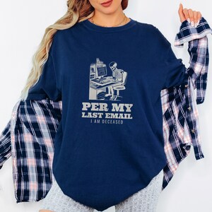 True Navy Comfort Colors t-shirt printed with the words Per My Last Email I Am Deceased and a drawing of a skeleton sitting at an office desk typing on a computer keyboard, worn by a woman