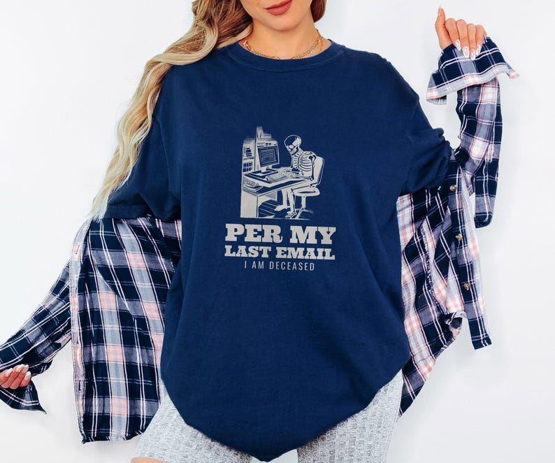 True Navy Comfort Colors t-shirt printed with the words Per My Last Email I Am Deceased and a drawing of a skeleton sitting at an office desk typing on a computer keyboard, worn by a woman