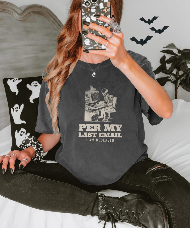 Pepper Comfort Colors t-shirt printed with the words Per My Last Email I Am Deceased and a drawing of a skeleton sitting at an office desk typing on a computer keyboard, worn by a woman