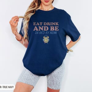 True Navy Comfort Colors t-shirt printed with the words Eat Drink And Be In Bed By Nine and a drawing of a retro brass wind-up alarm clock with its bells wrapped in fuzzy winter earmuffs, worn by a woman