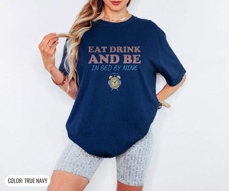 True Navy Comfort Colors t-shirt printed with the words Eat Drink And Be In Bed By Nine and a drawing of a retro brass wind-up alarm clock with its bells wrapped in fuzzy winter earmuffs, worn by a woman