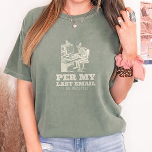 Moss Comfort Colors t-shirt printed with the words Per My Last Email I Am Deceased and a drawing of a skeleton sitting at an office desk typing on a computer keyboard, worn by a woman