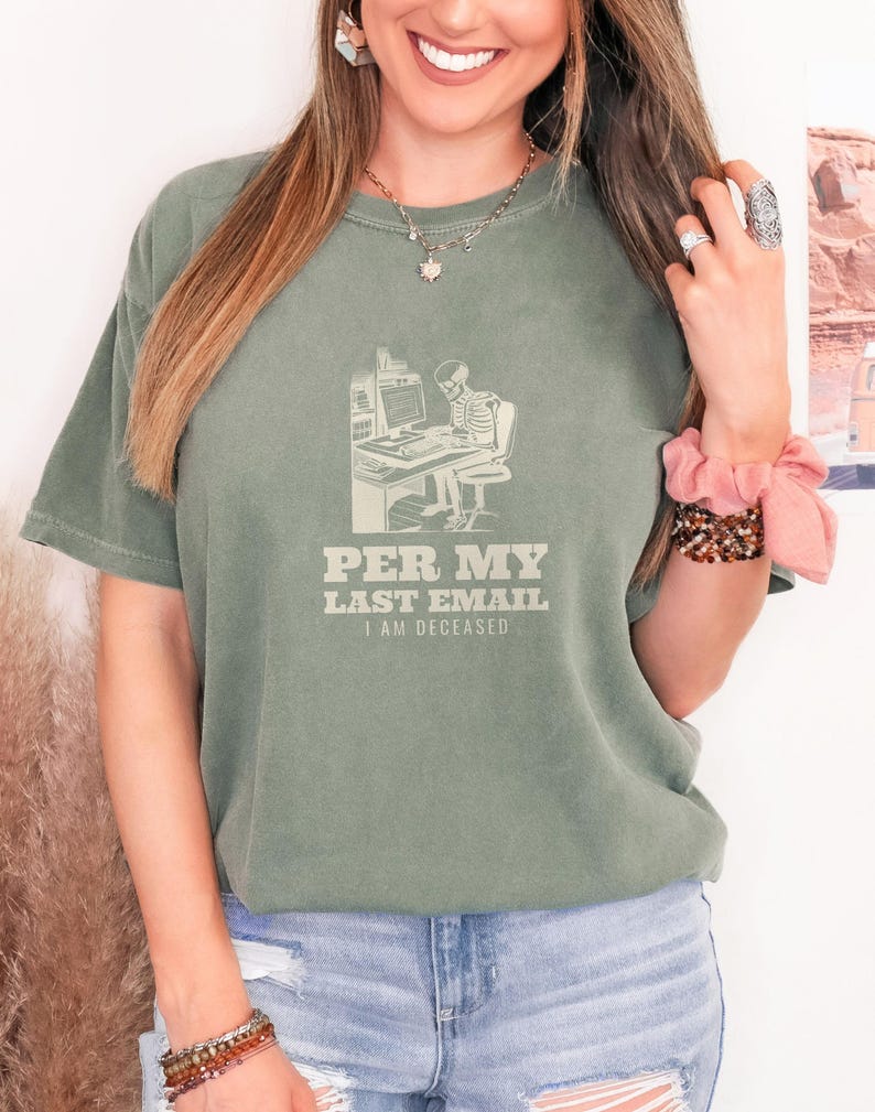 Moss Comfort Colors t-shirt printed with the words Per My Last Email I Am Deceased and a drawing of a skeleton sitting at an office desk typing on a computer keyboard, worn by a woman