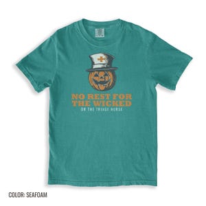 Seafoam Comfort Colors t-shirt printed with the words No Rest For The Wicked Or The Triage Nurse and a drawing of a grinning jack-o'-lantern wearing a classic nurse's cap with a red cross, folded flat lay