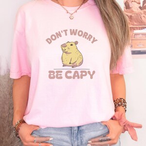 Blossom Comfort Colors t-shirt printed with the words Don'T Worry Be Capy and a drawing of a happy capybara with a small orange citrus slice on its head beside a green lily pad, worn by a woman