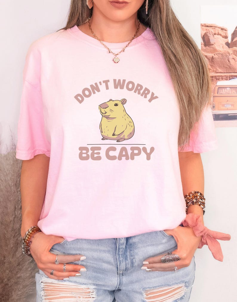 Blossom Comfort Colors t-shirt printed with the words Don'T Worry Be Capy and a drawing of a happy capybara with a small orange citrus slice on its head beside a green lily pad, worn by a woman