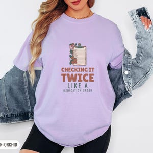 Orchid Comfort Colors t-shirt printed with the words Checking It Twice Like A Medication Order and a drawing of a classic wooden medical clipboard holding a paper medication checklist with checkboxes, a, worn by a woman