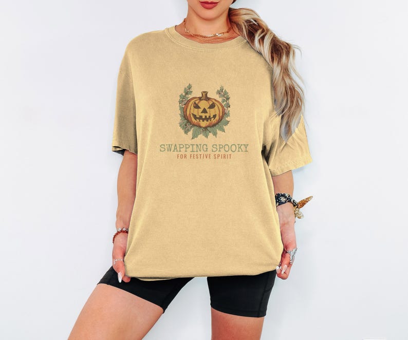 Butter Comfort Colors t-shirt printed with the words Swapping Spooky For Festive Spirit and a drawing of a spooky jack-o-lantern pumpkin alongside a festive holiday wreath, worn by a woman
