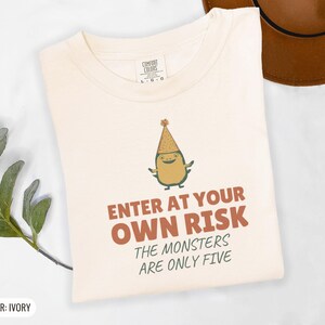 Ivory Comfort Colors t-shirt printed with the words Enter At Your Own Risk The Monsters Are Only Five and a drawing of a cute, smiling little green monster wearing a tiny paper party hat and holding a crayon, folded flat lay