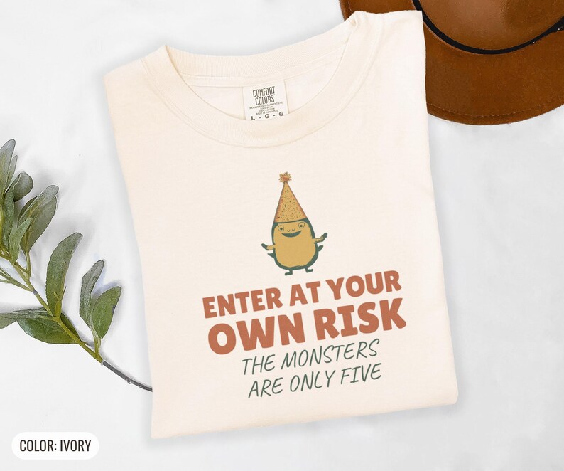 Ivory Comfort Colors t-shirt printed with the words Enter At Your Own Risk The Monsters Are Only Five and a drawing of a cute, smiling little green monster wearing a tiny paper party hat and holding a crayon, folded flat lay