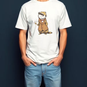 May include: A white short-sleeved t-shirt featuring a graphic illustration of a badger wearing a brown patterned vest and a bow tie, holding a small white teacup. The badger is depicted in a standing position with a tan and dark brown coat.