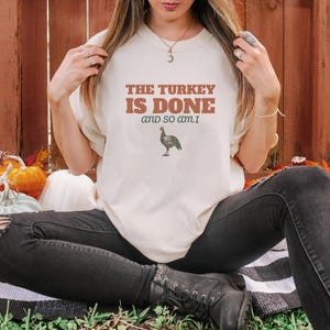 Ivory Comfort Colors t-shirt printed with the words The Turkey Is Done And So Am I and a drawing of a pop-up turkey timer stem popped up out of a roasted poultry leg, worn by a woman