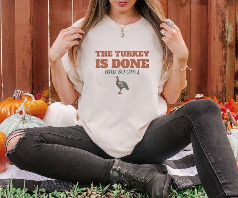 Ivory Comfort Colors t-shirt printed with the words The Turkey Is Done And So Am I and a drawing of a pop-up turkey timer stem popped up out of a roasted poultry leg, worn by a woman