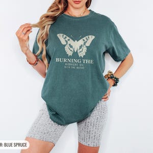 Blue Spruce Comfort Colors t-shirt printed with the words Burning The Midnight Oil With The Moths and a drawing of a cecropia moth with feathery antennae, worn by a woman