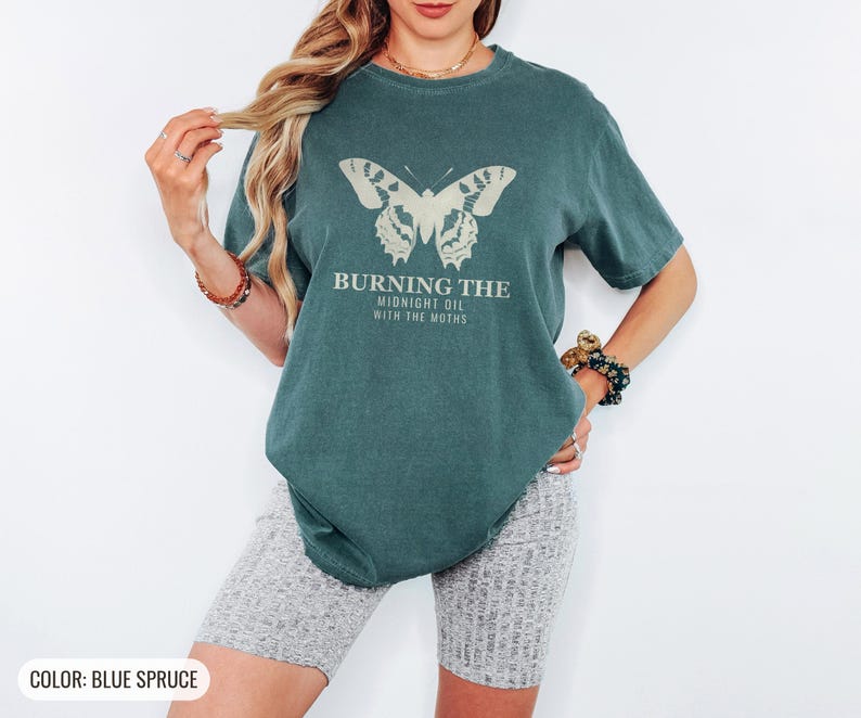 Blue Spruce Comfort Colors t-shirt printed with the words Burning The Midnight Oil With The Moths and a drawing of a cecropia moth with feathery antennae, worn by a woman