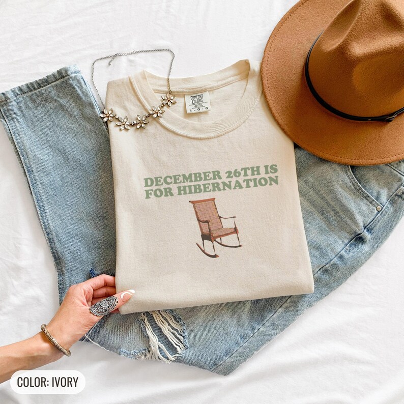 Ivory Comfort Colors t-shirt printed with the words December 26Th Is For Hibernation and a drawing of a cozy plaid wool blanket folded neatly on top of an oak rocking chair, folded flat lay