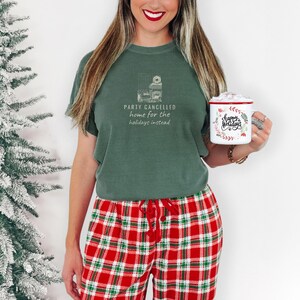 Moss Comfort Colors t-shirt printed with the words Party Cancelled Home For The Holidays Instead and a drawing of a cozy living room with a fireplace and Christmas decorations, a warm glow fills the space, worn by a woman