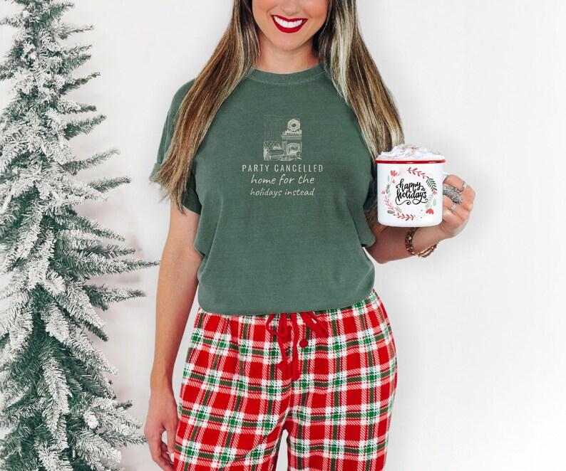 Moss Comfort Colors t-shirt printed with the words Party Cancelled Home For The Holidays Instead and a drawing of a cozy living room with a fireplace and Christmas decorations, a warm glow fills the space, worn by a woman