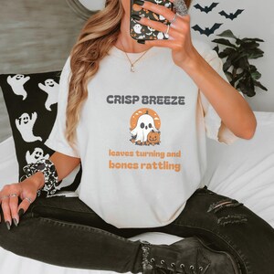 Ivory Comfort Colors t-shirt printed with the words Crisp Breeze Leaves Turning And Bones Rattling and a drawing of a large cute smiling ghost beside a carved jack-o-lantern, worn by a woman