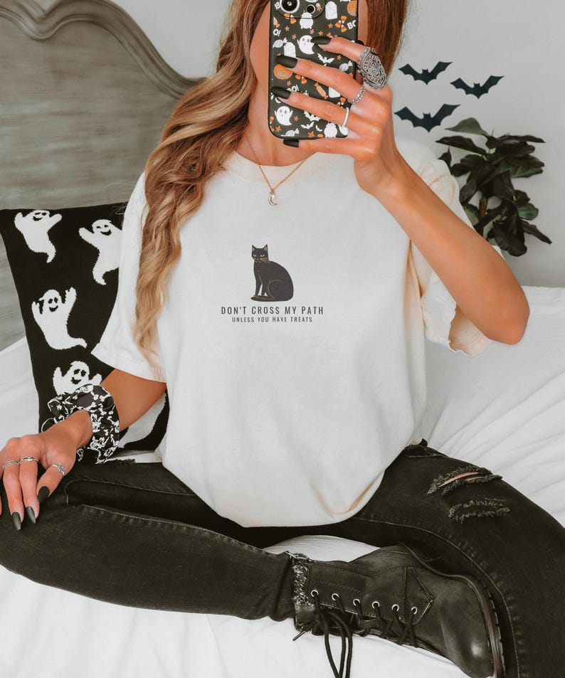 Ivory Comfort Colors t-shirt printed with the words Don'T Cross My Path Unless You Have Treats and a drawing of a cozy black cat wearing a tiny carved pumpkin collar, sitting in a stubborn pose, worn by a woman