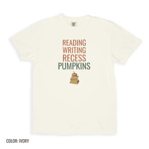 Ivory Comfort Colors t-shirt printed with the words Reading Writing Recess Pumpkins and a drawing of a stack of three vintage schoolbooks with a small orange pumpkin sitting on top, folded flat lay