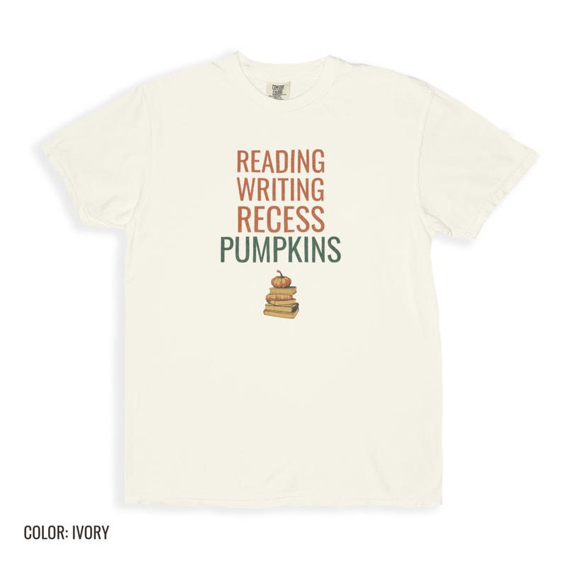 Ivory Comfort Colors t-shirt printed with the words Reading Writing Recess Pumpkins and a drawing of a stack of three vintage schoolbooks with a small orange pumpkin sitting on top, folded flat lay