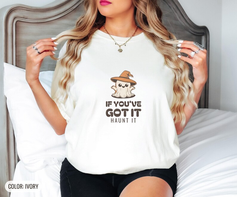Ivory Comfort Colors t-shirt printed with the words If You'Ve Got It Haunt It and a drawing of a cute retro cartoon ghost wearing a witch hat, worn by a woman